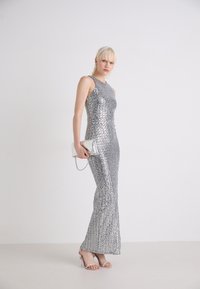Robe de soirée en sequins argentés avec un design ajusté, une épaule dénudée et un bas évasé. Associée à une pochette argentée et des talons à lanières.