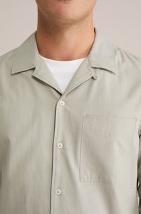 Kratke rokave, svetlozeleno srajco z ravnim ovratnikom, dvema gumboma in žepom na prsih. Narejena iz bombaža z gladko teksturo.