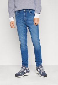 Jean en denim bleu clair avec une coupe slim, comportant cinq poches et une fermeture éclair. Associé à des baskets de sport grises et navy.