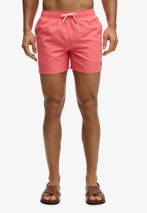 Mand iført koralrosa badeshorts med hvid snor og brune lædersandaler, stående mod en hvid baggrund.