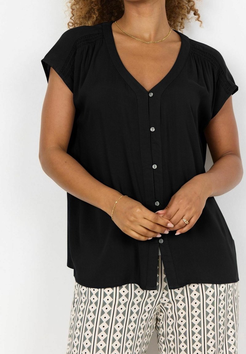 Blouse noire à boutons avec manches courtes, col en V, accents texturés sur les épaules et tissu doux. Portée avec un pantalon clair à motifs.