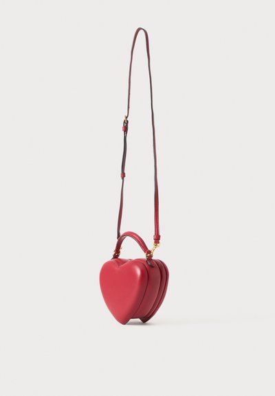 Sac à main en cuir rouge en forme de cœur avec poignée supérieure et longue bandoulière ajustable, présenté sur un fond uni clair.