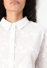 White embroidered blouse with a collared neckline, featuring floral patterns and button closures. Lightweight and textured fabric.