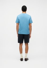 Homme debout, de dos, portant un t-shirt bleu clair, un short noir, des chaussettes crème et des chaussures habillées marron foncé sur un fond blanc uni.