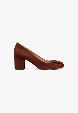 Scarpa in suede marrone con tacco a blocco, punta tondeggiante e dettaglio a fiocco sul davanti. Texture liscia con una leggera lucentezza e una struttura robusta.
