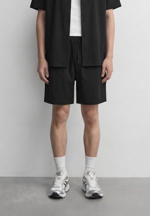 Person iført svarte shorts, svart kortermet skjorte med knapper over hvit T-skjorte, hvite ankelsokker og hvite løpesko.