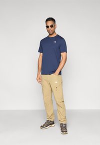 Πουκάμισο σε ναυτικό μπλε με λογότυπο, παντελόνι cargo σε ανοιχτό καφέ με φερμουάρ και μαύρα παπούτσια πεζοπορίας. Φορά γυαλιά ηλίου· απλή, λειτουργική ένδυση για υπαίθριες δραστηριότητες.