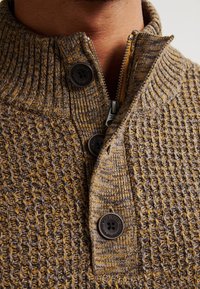 Close-up van een bruin en geel gebreid sweater met drie zwarte knopen en een gedeeltelijk ritssluiting kraag om iemands nek.