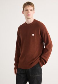 Baksteenrode, gestructureerde gebreide trui met ronde hals en geribde manchetten. Heeft een klein logo-opdruk op de linkerborst. Draagt het met donkere jeans.