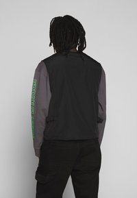 Chaleco negro sobre una camiseta de manga larga gris, con texto verde en la manga, con correas ajustables a los lados y una textura suave.