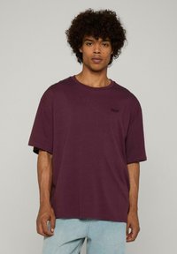 Túlméretezett, rövid ujjú póló sötét burgundi pamutból, kerek nyakkivágással és kis fekete szöveges részlettel a mellrészen. Világoskék farmernadrággal párosítva.