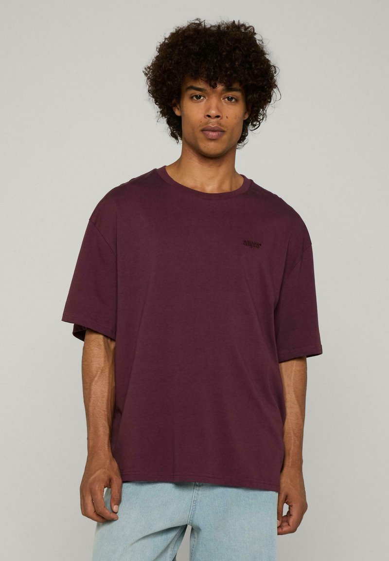 Túlméretezett, rövid ujjú póló sötét burgundi pamutból, kerek nyakkivágással és kis fekete szöveges részlettel a mellrészen. Világoskék farmernadrággal párosítva.