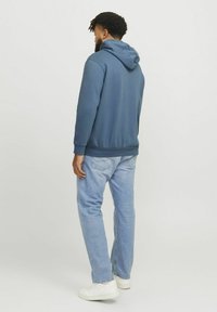Hættebluse i blåt stof med en glat tekstur, parret med lyseblå jeans og hvide sneakers, set fra ryggen.