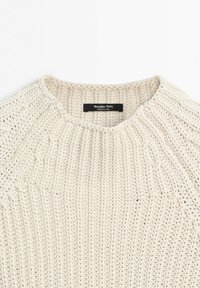 Kremowy, gruby sweter dziany o okrągłym dekolcie z widocznymi wzorami splotu prążkowanego i zaplatanego.