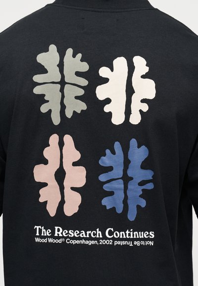 Μαύρο πίσω μέρος πουκάμισου με τέσσερα αφηρημένα σχήματα φύλλων σε γκρι, άσπρο, ροζ και μπλε πάνω από το κείμενο που γράφει «Η Έρευνα Συνεχίζεται» και λεπτομέρειες της μάρκας.