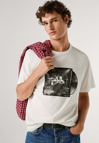 Witte katoenen t-shirt met een drummende afbeelding en een vinylplatenontwerp. Model draagt een rood geruit hemd over de schouder, gecombineerd met een spijkerbroek.