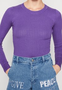 Persona che indossa un top lungo aderente e a coste di colore viola infilato in un paio di jeans blu a vita alta, con le scritte "GIVE" e "PEACE" ricamate sulle tasche frontali.
