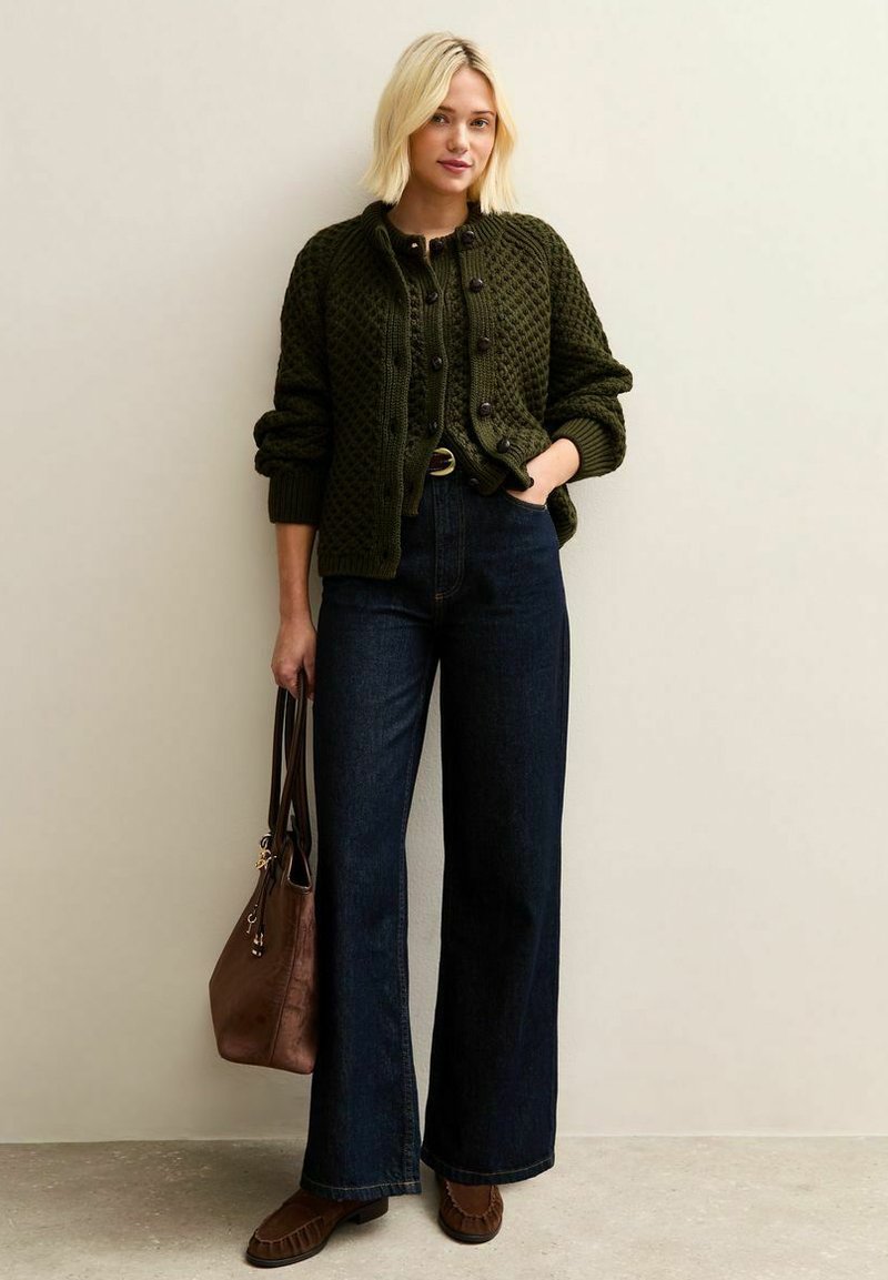Groene gebreide cardigan met diamantenpatroon, donkerblauwe wijde jeans, bruine leren tas en bijpassende schoenen. De knopen op de cardigan zijn opvallend.