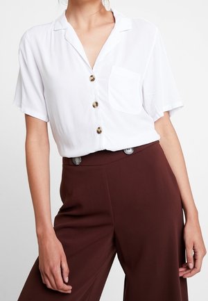 Vrouw draagt een wit, korte mouwen, knoopblouse met een borstzak en hoog getailleerde, donkerbruine wijde broek.