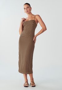 Žena v obtiahnutých, bezrukávových midi šatách s hnedým a čiernym geometrickým vzorom, so zlatými kruhovými náušnicami a čiernymi sťahovacími sandálmi.