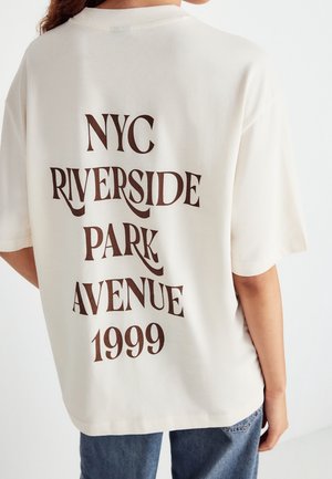 Person iført offwhite t-shirt med "NYC Riverside Park Avenue 1999" i store brune bogstaver på ryggen, kombineret med blå jeans.