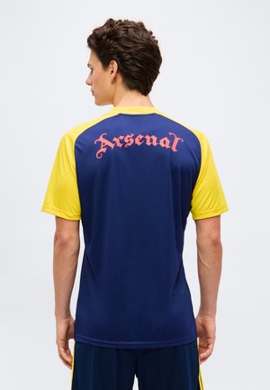 Mann trägt ein marineblaues und gelbes Sportshirt mit „Arsenal“ in roter gotischer Schrift auf dem Rücken und steht vor einem schlichten hellen Hintergrund.