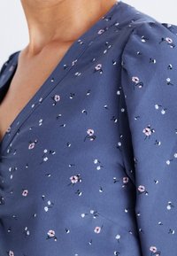 Gros plan sur une personne portant un chemisier bleu avec de petites imprimés floraux roses et blancs et un décolleté en V.