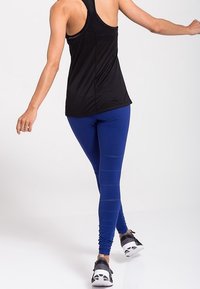 Černý sportovní nátělník bez rukávů, modré legíny s texturovanými pruhy a šedé sportovní boty s černými akcenty. Modelka je zobrazena zezadu.