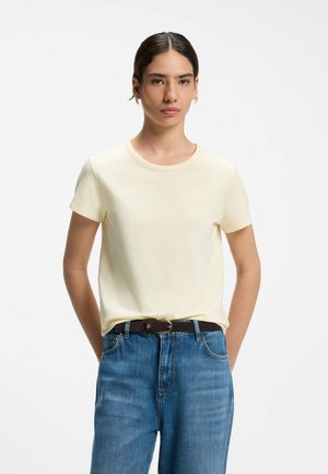 Donna con capelli scuri con una t-shirt crema a maniche corte infilata in jeans blu con cintura nera, in piedi davanti a uno sfondo bianco uniforme.