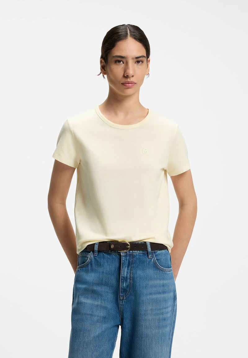 Femme aux cheveux foncés portant un t-shirt crème à manches courtes, rentré dans un jean bleu avec une ceinture noire, debout devant un fond blanc uni.