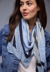 Jeune femme aux longs cheveux bruns portant une écharpe bleue à motifs et une veste en jean, regardant légèrement de côté sur un fond gris.