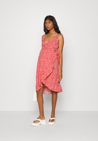 Vestido vermelho envolvente com pequeno padrão floral branco, decote em V e barra com babados. Modelo está usando sandálias brancas de plataforma, posando contra um fundo neutro.