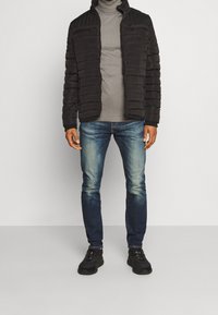 Svart puffjacka med horisontellt kviltat mönster, grå polotröja, blå denimjeans och svarta sneakers. Enkel, funktionell design.