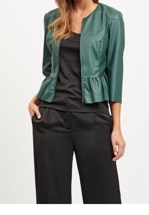 Femme portant une veste peplum vert foncé à manches trois-quarts sur un haut noir et un pantalon noir à jambes évasées.