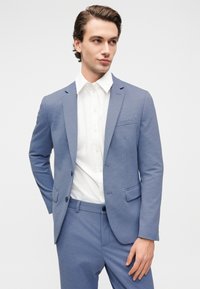 Ανοιχτό μπλε, υφασμάτινο κοστούμι με σακάκι μονόμπουμπο και ταιριαστό παντελόνι. Συνδυάζεται με λευκό πουκάμισο με κουμπιά. Απλός και μοντέρνος σχεδιασμός.