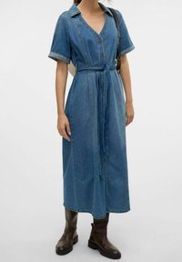 Jeans-Midi-Kleid in Hellblau mit kurzen Ärmeln, V-Ausschnitt, Vorderknöpfen und einem Bindegürtel. Getragen mit braunen Knöchelschuhen.