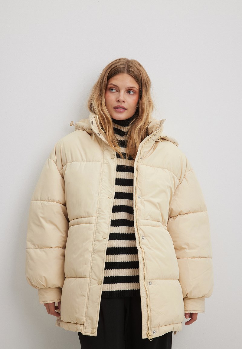 Béžová prešívaná bunda s kapucňou, cvokmi a prešívanou textúrou. Nosená cez čierno-biely pruhovaný sveter. Dlhé rukávy.