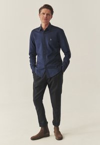 Tumši zils krekls ar pogām un mazu logotipu, savienots ar tumšiem biksēm un brūniem zamšādās zābakiem. Krekls ir ar klasisku apkakli un garām piedurknēm.