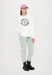 Felpa bianca con grafica floreale e di farfalla, abbinata a jeans azzurri chiari e stivali militari neri. Cappellino rosa indossato al contrario.