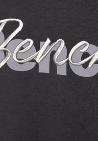 Dunkelgrauer Stoff mit einem auffälligen silbernen gestickten Schriftzug "Bene". Die Textur ist glatt mit einem leichten Schimmer.