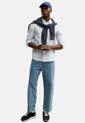 Man met lichtblauw gestreept overhemd met knopen, een donkerkleurige trui over de schouders gedrapeerd, blauwe spijkerbroek, zwarte sneakers en een blauwe honkbalpet.