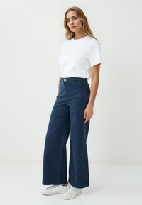 Szerokie granatowe spodnie z wysokim stanem, wykonane z gładkiego materiału. Posiadają zapięcie na guzik z przodu oraz wyraźne szwy. W zestawie z białą koszulką.