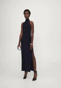 Sieviete, tērpusies melnā bezgarša halter-neck kleitā ar augstu šķēlumu, melnās augstpapēžu kurpēs, ar sudraba aproci un turot sudraba rokassomiņu.