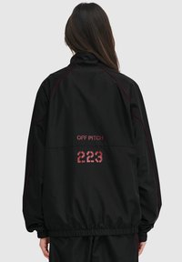 Sort vindjakke med røde accenter, der har høj krave, elastiske manchetter og trykte tekster "OFF PITCH" og "223" på ryggen.