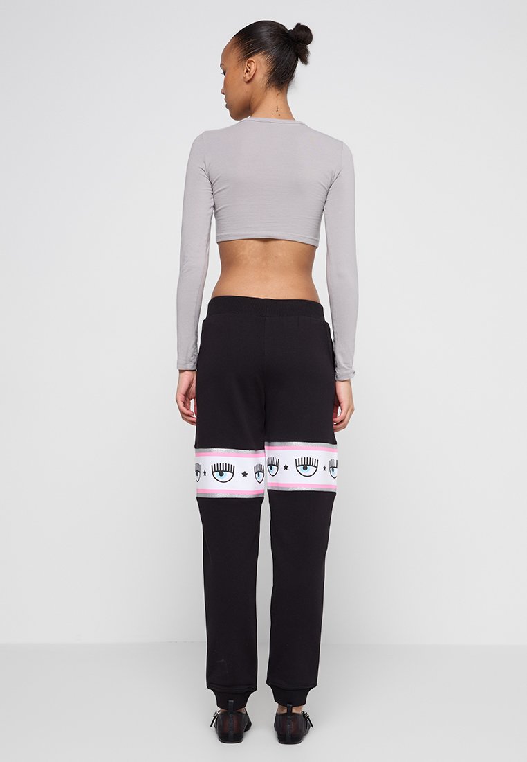 Schwarze Sweatpants mit einem pinken Grafikband, das Augenmotive zeigt; kombiniert mit einem enganliegenden grauen Langarmoberteil im verkürzten Design.