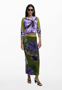 Gebreide trui in groen en paars bloemenpatroon; gecombineerd met een lange groene rok met soortgelijke bloemenontwerpen. Model draagt zwarte zonnebril en sandalen.