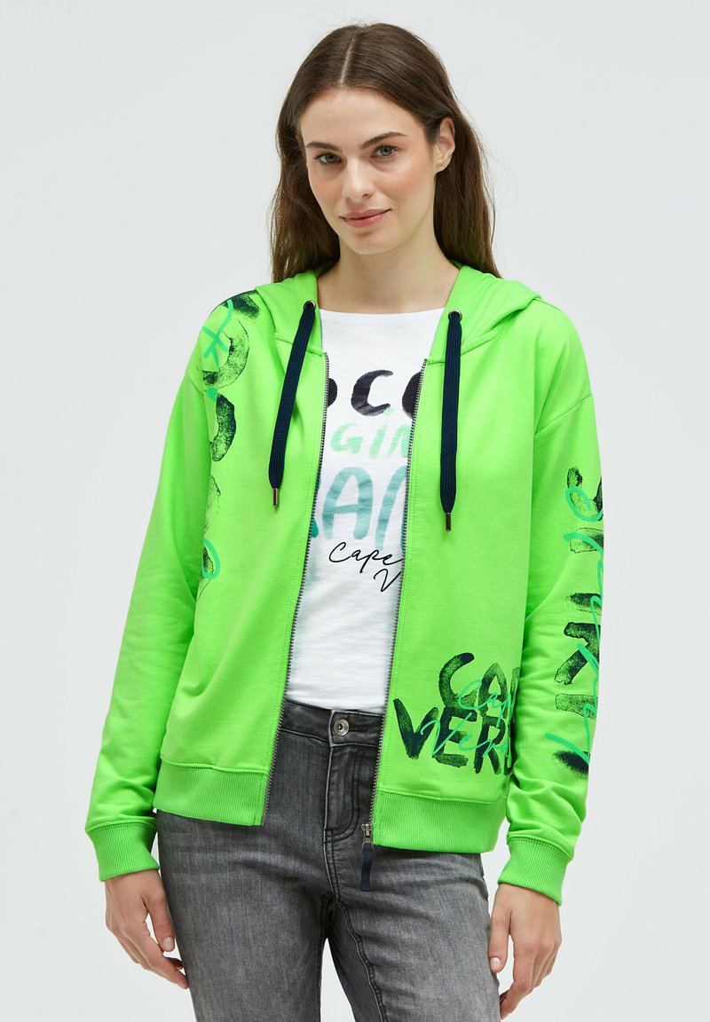 Junge Frau trägt eine leuchtend grüne Kapuzenjacke mit Reißverschluss, schwarzen Kordeln und schwarzem Text auf den Ärmeln, kombiniert mit grauen Jeans und einem weißen, bedruckten T-Shirt.