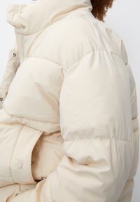 Giacca beige puffosa con colletto alto, dotata di bottoni a pressione e tessuto testurizzato. Il design include maniche voluminose e una tasca laterale.
