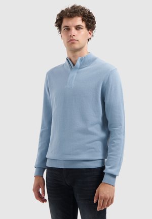 Jonge man met krullend haar, gekleed in een lichtblauwe trui met hoge kraag en zwarte broek, staand tegen een effen achtergrond.