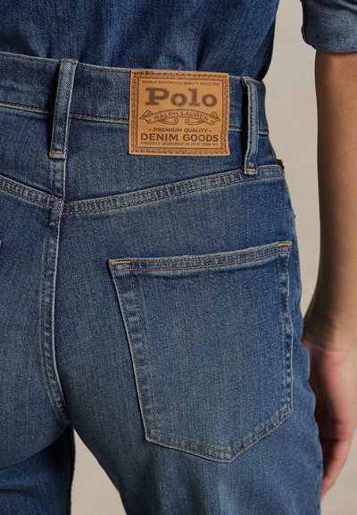 Mėlynų džinsų užpakalinės kišenės ir liemens artimas planas su šviesiai rudos odos Polo Ralph Lauren prekės ženklo etikete.